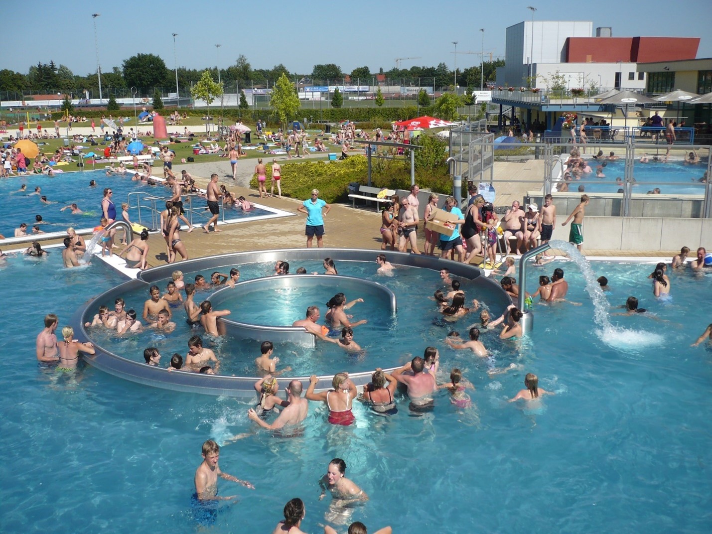 Ein weitläufiger Pool, gefüllt mit Menschen, die schwimmen und sich amüsieren, umgeben von Sonnenlicht und entspannter Atmosphäre.