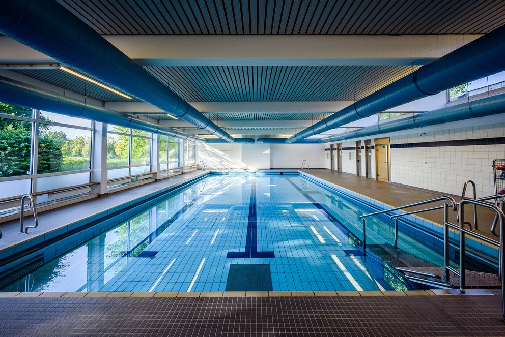 Lichtdurchflutetes Hallenbad mit Wettkampfbecken Modernes Hallenbad mit einem langen Schwimmbecken, blauen Bahnmarkierungen und großen Fenstern, durch die Tageslicht hereinfällt. Die Decke ist mit einem auffälligen Muster versehen, und am Beckenrand stehen Sitzgelegenheiten und Schwimmausrüstung