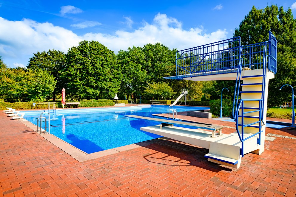 Außenbecken mit Sprunganlage. Das Bild zeigt ein Freibad mit Sprungturm vor grünem Baumbestand. Das klare Wasser, die rote Pflasterung und der blaue Himmel schaffen eine einladende Atmosphäre für Sommer, Spaß und sportliche Aktivitäten.