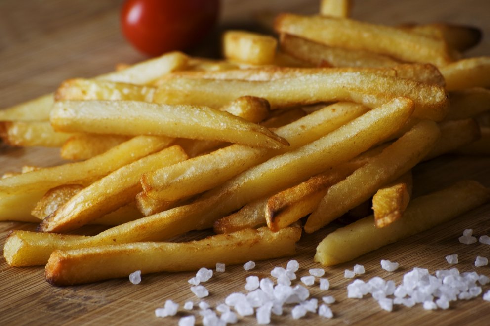 Es liegen Pommes ohne Teller auf dem Holztisch, Salzkörner im Vordergrund. Im Hintergrund ist eine Tomate zu sehen