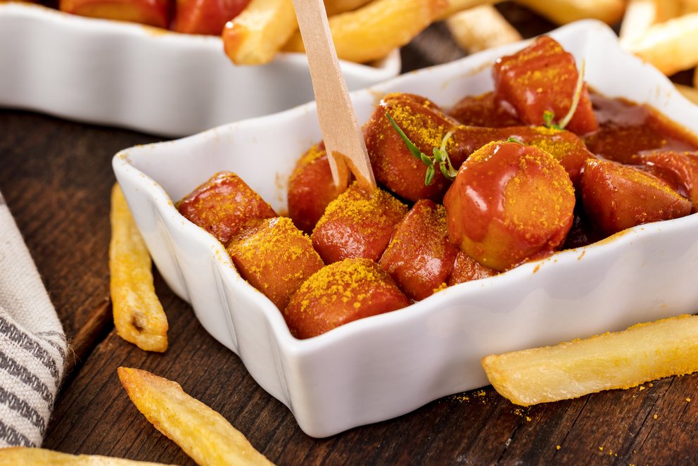 Eine Currywurst in einer Schale, diese steht auf einem Holztisch. Es liegen Pommes drum herum