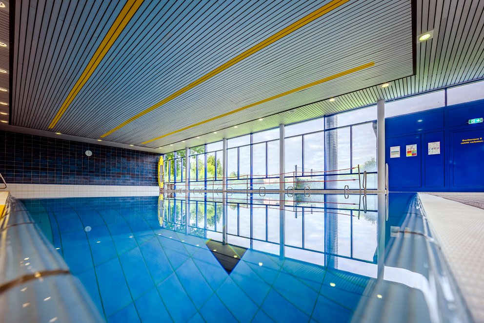 Innenpool mit blauer Decke und lichtdurchflutetem Ambiente Schwimmbad in Innenräumen mit einer blauen Decke, die eine entspannende Atmosphäre schafft, umgeben von hellen Wänden.